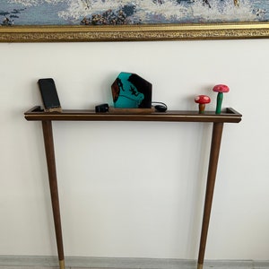 Puede incluir: Una mesa consola de madera marr&oacute;n oscuro con detalles dorados y dos patas. La mesa tiene una superficie rectangular con una escultura de resina turquesa y dos figuritas de setas rojas y blancas en la parte superior.