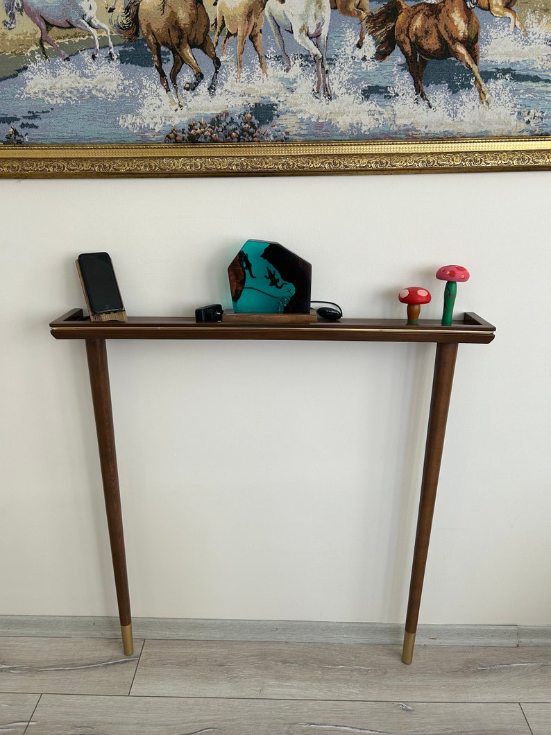 Puede incluir: Una mesa consola de madera marr&oacute;n oscuro con detalles dorados y dos patas. La mesa tiene una superficie rectangular con una escultura de resina turquesa y dos figuritas de setas rojas y blancas en la parte superior.