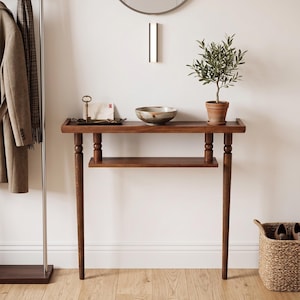 May include: A wooden entryway table with a lower shelf and four tapered legs. A small potted olive tree sits on the table, along with a decorative bowl and a tray with a key and mail. A coat rack and a woven basket are also visible.