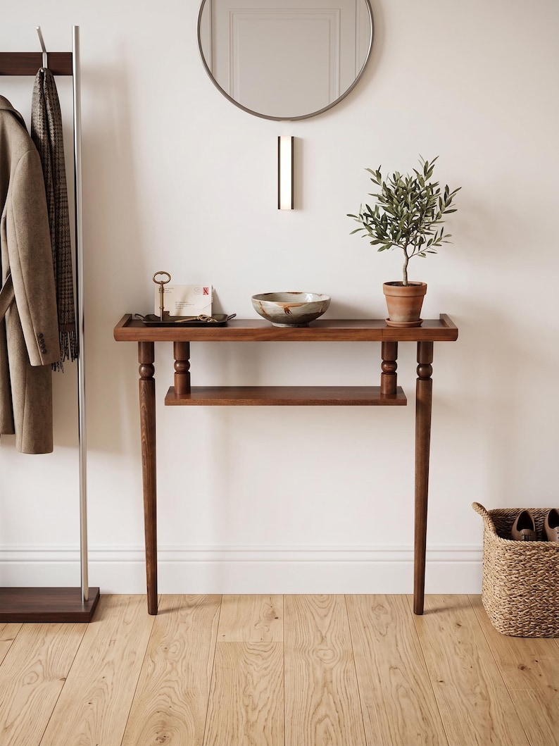 May include: A wooden entryway table with a lower shelf and four tapered legs. A small potted olive tree sits on the table, along with a decorative bowl and a tray with a key and mail. A coat rack and a woven basket are also visible.