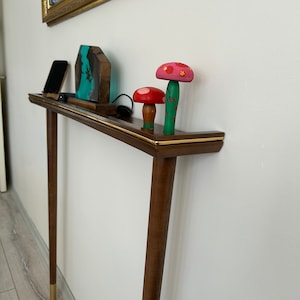 Puede incluir: Una consola de madera con una tapa marr&oacute;n y un borde dorado. La mesa tiene dos patas y est&aacute; decorada con una peque&ntilde;a escultura de resina azul y verde, un tel&eacute;fono m&oacute;vil negro y tres figuritas de setas de madera. Las setas est&aacute;n pintadas en tonos de rosa, rojo y verde.