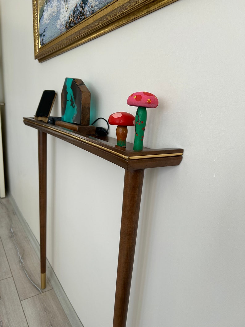 Puede incluir: Una consola de madera con una tapa marr&oacute;n y un borde dorado. La mesa tiene dos patas y est&aacute; decorada con una peque&ntilde;a escultura de resina azul y verde, un tel&eacute;fono m&oacute;vil negro y tres figuritas de setas de madera. Las setas est&aacute;n pintadas en tonos de rosa, rojo y verde.