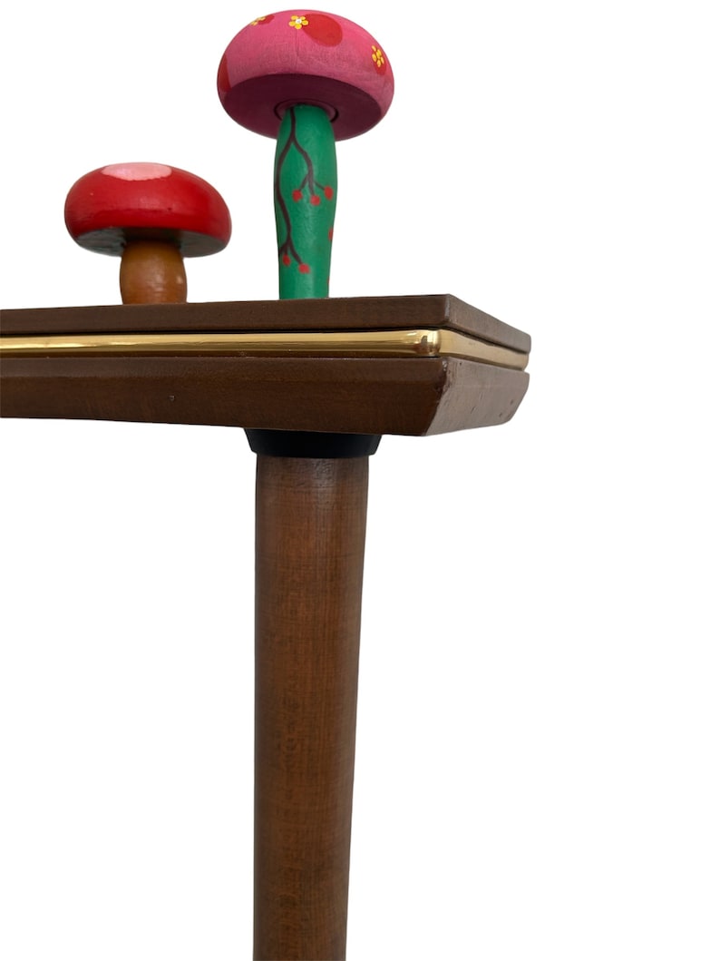 Puede incluir: Una mesa de madera con un borde dorado y una pata de madera marr&oacute;n. Dos figuritas de hongos de madera est&aacute;n encima de la mesa. Un hongo es rosa con flores amarillas y el otro es rojo con puntos blancos.