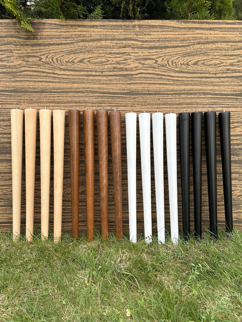 May include: A set of 12 wooden table legs in various colors, including natural wood, brown, white, and black. The legs are all the same size and shape, and they are arranged in rows on a wooden surface.