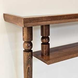 May include: A close-up of a wooden console table with a dark brown finish. The table features a rectangular top, a lower shelf, and turned legs with a ball and cylinder design. The wood grain is visible.
