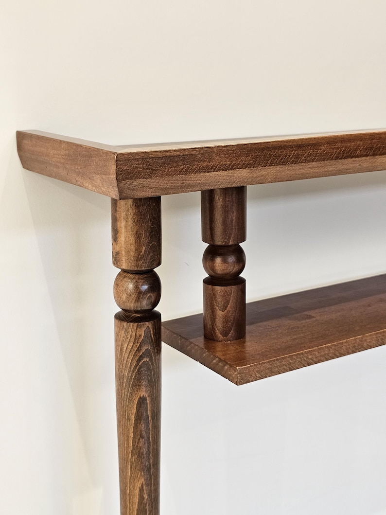 May include: A close-up of a wooden console table with a dark brown finish. The table features a rectangular top, a lower shelf, and turned legs with a ball and cylinder design. The wood grain is visible.