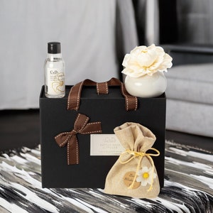 May include: A black gift box with a brown ribbon and bow. The box contains a clear bottle of essential oil, a white ceramic diffuser with a white flower, and a small hessian bag with a tag that says "Best Wishes".