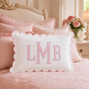 May include: A rectangular white throw pillow featuring a scalloped edge trimmed in light pink. The center of the pillow displays a large, embroidered monogram with the letters LMB in a light pink font with a light blue outline.