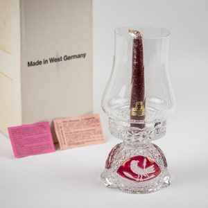 May include: A clear glass candle holder with a red base and a cut crystal design. The holder features a bird with a wheat stalk in its beak. The candle holder is made in West Germany.