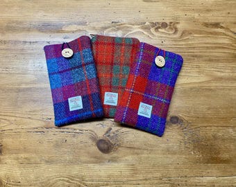 May include: Three plaid fabric cases in red, blue, and green, each with a wooden button fastening. The cases are arranged on a wooden surface. Each case has a small white label with text.