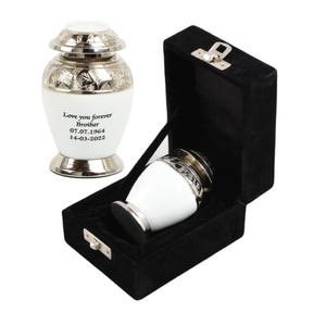 May include: White cremation urn with silver trim and engraved inscription "Love you forever Brother 07.07.1964 14-03-2022". The urn is in a black velvet box with a silver latch.