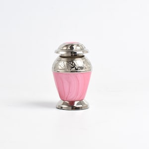 May include: A small pink cremation urn with silver accents and a floral design. The urn has a lid and is made of metal.