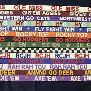 May include: A collection of colorful fabric straps with various college team slogans, including "Ole Miss", "Gig'em Aggies", "Northwestern Go 'Cats", "Fly Fight Win", "Rocky Top", "Go Hoosiers", "Sic 'Em", "RCJH", "Rock Chalk", "Rah Rah TCU", "Annnd Go Deer", and "Axe 'Em".