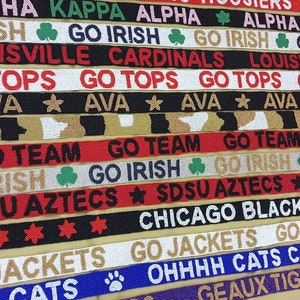 May include: A collection of colorful beaded straps with various college and team logos and slogans, including "Go Irish", "Go Tops", "Go Team", "SDSU Aztecs", "Chicago Blackhawks", "Go Jackets", "Ohhhhh Cats", and "Geaux Tigers".