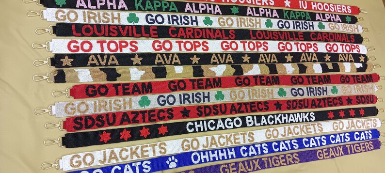 May include: A collection of colorful beaded straps with various college and team logos and slogans, including "Go Irish", "Go Tops", "Go Team", "SDSU Aztecs", "Chicago Blackhawks", "Go Jackets", "Ohhhhh Cats", and "Geaux Tigers".