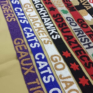 May include: A collection of beaded straps with various college and university team names and logos. The straps are made with white, black, red, blue, gold, and green beads. The straps are arranged in a row, with the names and logos facing the viewer.