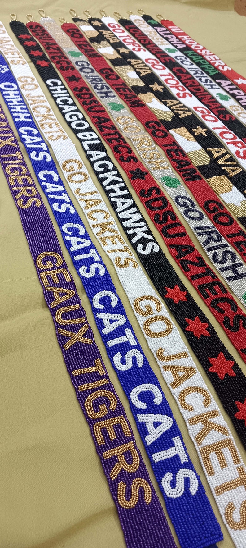 May include: A collection of beaded straps with various college and university team names and logos. The straps are made with white, black, red, blue, gold, and green beads. The straps are arranged in a row, with the names and logos facing the viewer.