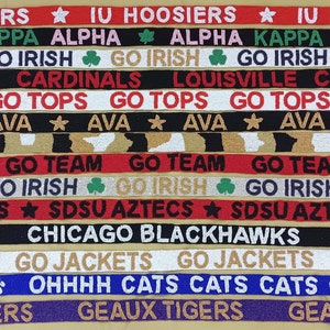May include: A collection of colorful fabric wristbands with various college and team logos and slogans, including "IU Hoosiers", "Go Irish", "Louisville Cardinals", "Go Tops", "Go Team", "SDSU Aztecs", "Chicago Blackhawks", "Go Jackets", "Go Cats", and "Geaux Tigers".