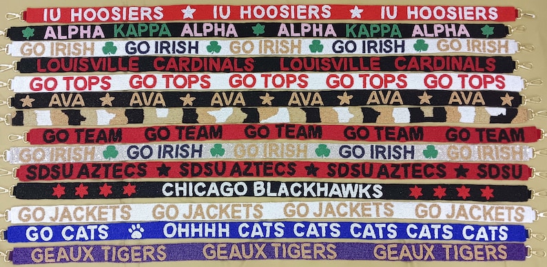 May include: A collection of colorful fabric wristbands with various college and team logos and slogans, including "IU Hoosiers", "Go Irish", "Louisville Cardinals", "Go Tops", "Go Team", "SDSU Aztecs", "Chicago Blackhawks", "Go Jackets", "Go Cats", and "Geaux Tigers".