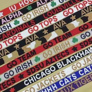 May include: A collection of colorful woven straps with various college team names and logos, including IU Hoosiers, Alpha Kappa Alpha, Go Irish, Louisville Cardinals, Go Tops, Ava, Go Team, SDSU Aztecs, Chicago Blackhawks, Go Jackets,  Go Cats, Geaux Tigers.