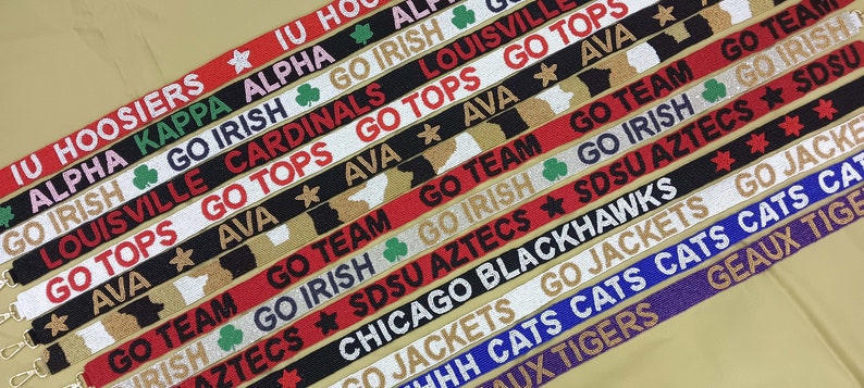 May include: A collection of colorful woven straps with various college team names and logos, including IU Hoosiers, Alpha Kappa Alpha, Go Irish, Louisville Cardinals, Go Tops, Ava, Go Team, SDSU Aztecs, Chicago Blackhawks, Go Jackets,  Go Cats, Geaux Tigers.