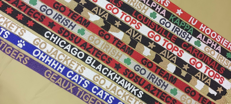 May include: A close-up of ten colorful beaded straps with various college team names and logos. The straps feature text such as "Go Irish", "Go Tops", "Go Team", "SDSU Aztecs", "Chicago Blackhawks", "Go Jackets", "Ohhhhh Cats", "Geaux Tigers", "IU Hoosier", "Kappa Alpha", "Louisville Cardinals", and "Go Irish".