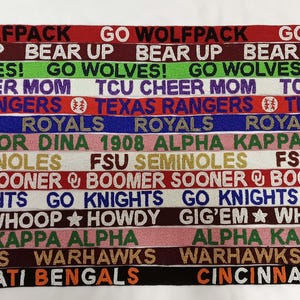 May include: A close-up of a collection of colorful woven wristbands with various team names and logos, including "Go Wolfpack", "Bear Up", "Go Wolves!", "TCU Cheer Mom", "Texas Rangers", "Royals", "Soror Dina 1908 Alpha Kappa Alpha", "FSU Seminoles", "Boomer Sooner", "Go Knights", "Gig'em Whoop Howdy", "Alpha Kappa Alpha", "Warhawks", and "Cincinnati Bengals".