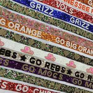 May include: A collection of colorful, glittery straps with various team names and slogans, including "Go Big Orange", "Go Vols", "Go Rebels", "Go Mocs", and "Go Chiefs".