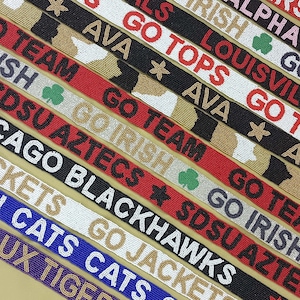 May include: A close-up of colorful fabric ribbon with various college team names and logos. The ribbons are black, white, red, gold, and blue with white lettering. The text includes "Go Irish", "Go Jackets", "Go Team", "SDSU Aztecs", "Go Tops", "Louisville Cardinals", "IU Hoosiers", "Kappa Alpha", "Chicago Blackhawks", "OHHHH Geaux Tigers", "Go Cats", and "Go Irish".