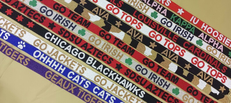 May include: A close-up of colorful fabric ribbon with various college team names and logos. The ribbons are black, white, red, gold, and blue with white lettering. The text includes "Go Irish", "Go Jackets", "Go Team", "SDSU Aztecs", "Go Tops", "Louisville Cardinals", "IU Hoosiers", "Kappa Alpha", "Chicago Blackhawks", "OHHHH Geaux Tigers", "Go Cats", and "Go Irish".
