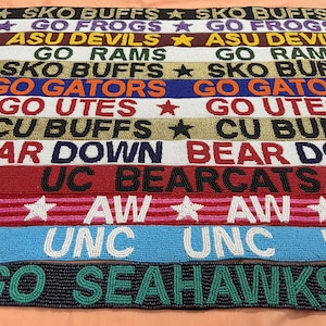 May include: A collection of beaded belts with various team names and logos, including "SKO BUFFS," "GO FROGS," and "GO GATORS." The belts feature a variety of colors, including black, purple, gold, blue, and red, with white star accents.
