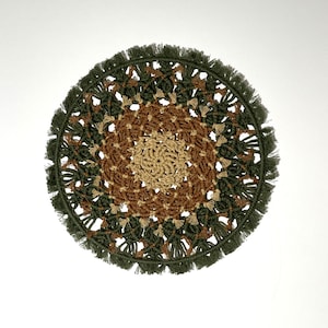 May include: A round macrame wall hanging with a green, brown, and beige color scheme. The hanging has a fringe border and a geometric pattern.