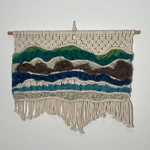 May include: A wall hanging made of macrame with a woven design featuring blue, green, and brown yarn. The hanging has a fringe border.