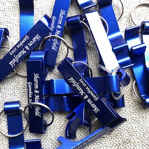 May include: A pile of blue metal bottle openers with key rings. Each opener is engraved with the text "Sharon & Mansfield" and the date "Gambia 2023".