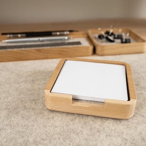 May include: A square light wood holder containing a stack of plain white square paper notes. In the background, a rectangular light wood desk organizer holds a black pen, a silver pen, a dark ruler, and a small square tray with black clips.
