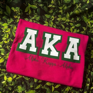 May include: A magenta t-shirt with the letters "AKA" in white block letters with green trim. Below the letters is the cursive text "Alpha Kappa Alpha". The shirt is displayed on a bed of green leaves.
