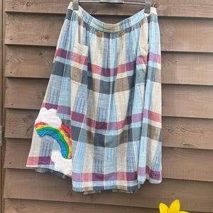 May include: A plaid skirt in shades of blue, purple, and brown, with a rainbow and cloud applique. The skirt is hanging on a hanger against a wooden background. The skirt's design features a pleated waist and a knee-length hemline.