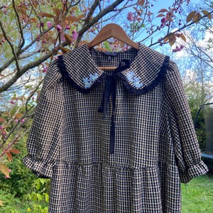 May include: A black and beige gingham dress with a large, frilled collar. The collar is trimmed with a navy ruffle and decorated with small blue floral embroidery. The dress has short sleeves and a navy ribbon tie.