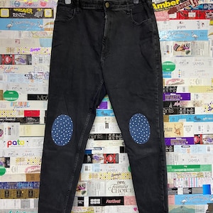 May include: Black denim jeans with blue polka dot knee patches. The jeans are hanging on a black hanger. The background is a collage of colorful labels and packaging.