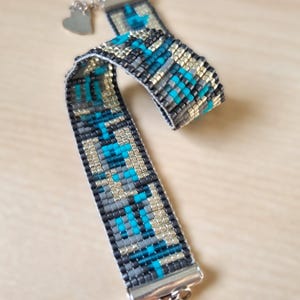 Puede incluir: Un brazalete de cuentas con un patrón geométrico en tonos grises, azules y dorados. El brazalete tiene un cierre plateado y un pequeño dije en forma de corazón.