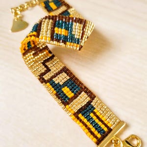 Puede incluir: Un brazalete de cuentas doradas con un patrón geométrico en marrón, amarillo y azul verdoso. El brazalete tiene un cierre en forma de corazón y un cierre de langosta.