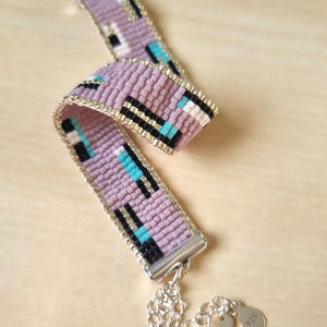 Könnte beinhalten: Ein Armband aus Perlen mit einem geometrischen Muster in Rosa-, Schwarz-, Weiß- und Türkistönen. Das Armband hat einen silbernen Verschluss und einen silbernen Anhänger in Herzform mit der Aufschrift "925".