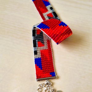 Könnte beinhalten: Ein Armband aus Perlen mit einem Muster aus roten, blauen, schwarzen und silbernen Perlen. Es hat einen silbernen Verschluss und einen herzförmigen Anhänger. Das Armband besteht aus kleinen, farbigen Perlen.