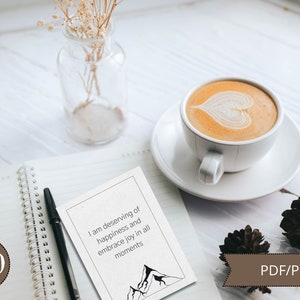 May include: A white card with a black mountain range illustration and the text "I am deserving of happiness and embrace joy in all moments". The card is on a white notebook with a black pen and a brown circle with the number 100.