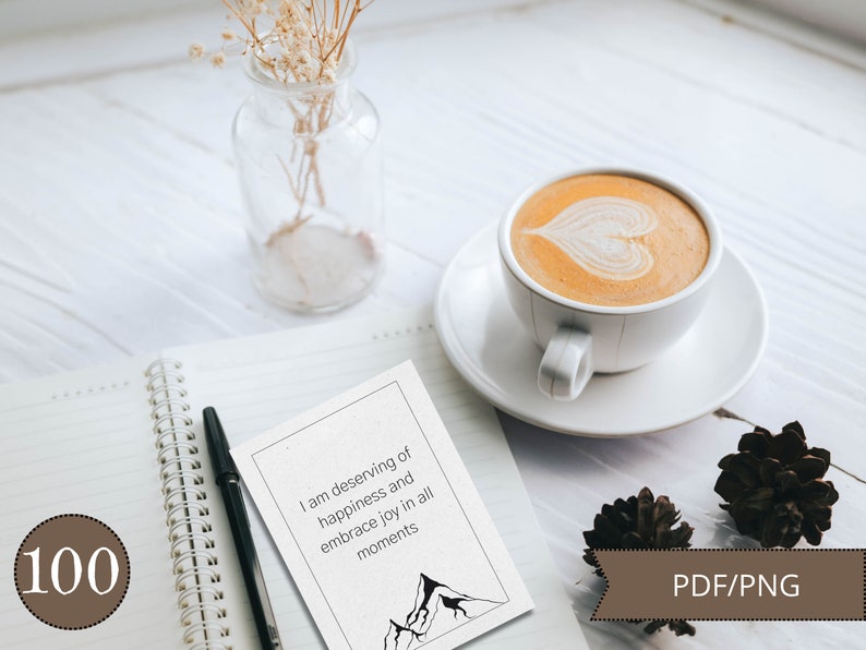 May include: A white card with a black mountain range illustration and the text "I am deserving of happiness and embrace joy in all moments". The card is on a white notebook with a black pen and a brown circle with the number 100.