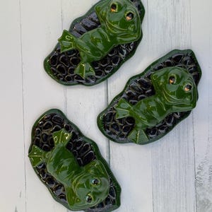 May include: Three glossy green ceramic frog figurines resting on lily pads. The frogs have detailed features and are positioned on textured, dark green and black lily pads. The background is a light wood grain.