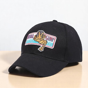 Könnte beinhalten: Schwarze Baseballkappe mit dem Logo "Bubba Gump Shrimp Co.". Das Logo ist ein rechteckiger Aufnäher mit einer Cartoon-Garnele und Text in Blau, Rot und Weiß. Die Kappe hat einen gebogenen Schirm und einen Knopf oben.