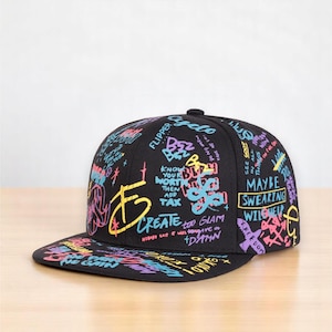 May include: Black flat-brimmed baseball cap with colorful graffiti-style text and designs. The text includes phrases like "Create," "Maybe Swearing," and "Freedom." The cap has a structured fit and a flat brim, suitable for casual wear.