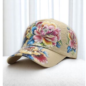 Pode incluir: Um boné de beisebol bege com estampa floral. As flores são rosa, amarelo, azul e verde. O boné tem uma parte de trás de malha.