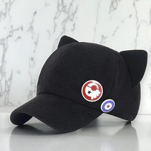 May include: Black baseball cap with cat ears. The cap features two round pins: one with a skull and star design, and another with a blue and white target symbol. The cap is made of a soft material.